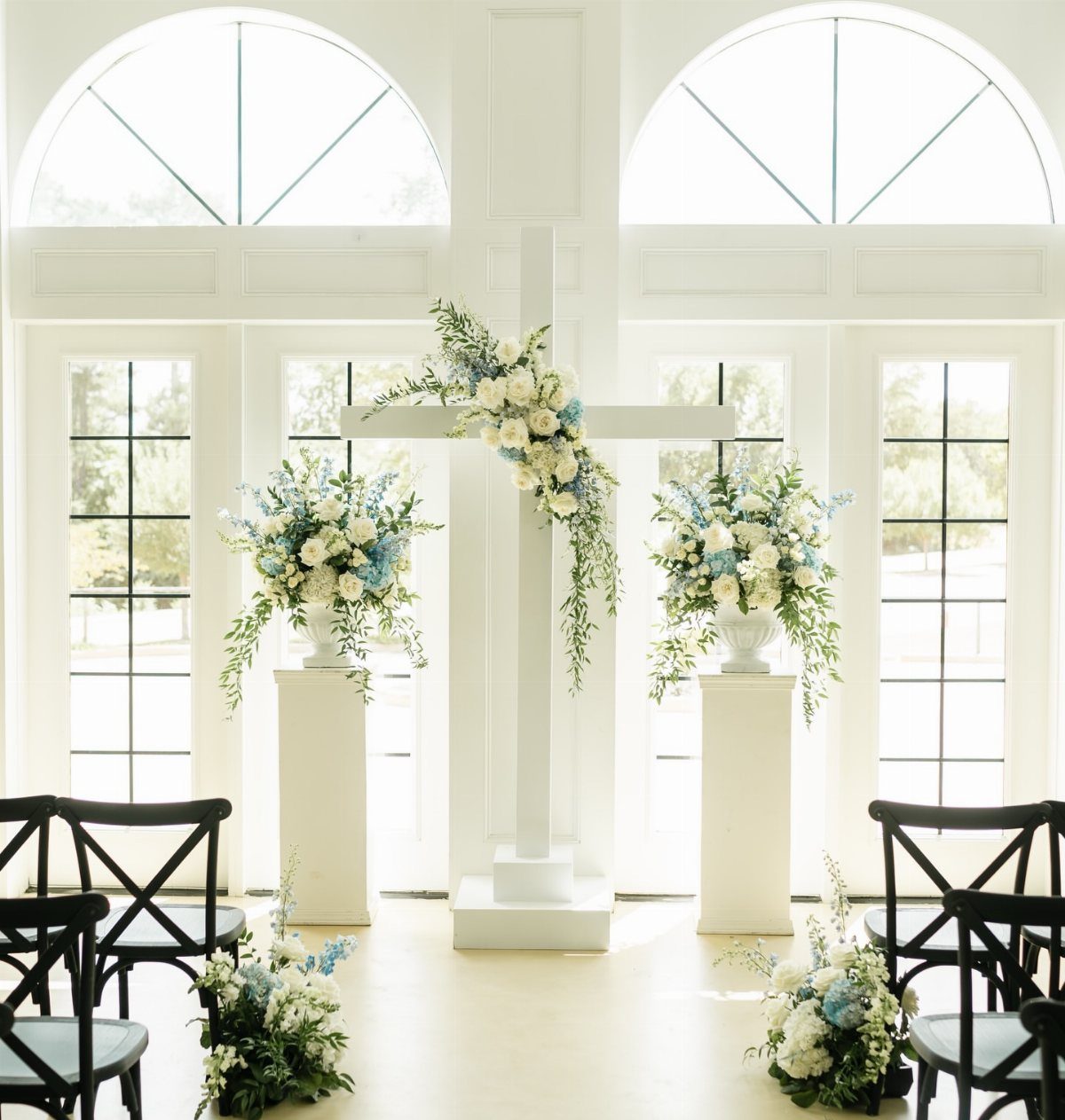 A large white wooden cross decorated with white and blue flowers in a bright room.