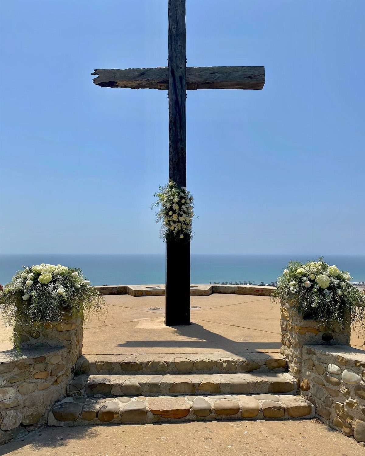 A large wooden cross decorated with white flowers stands on a stone terrace overlooking the ocean.