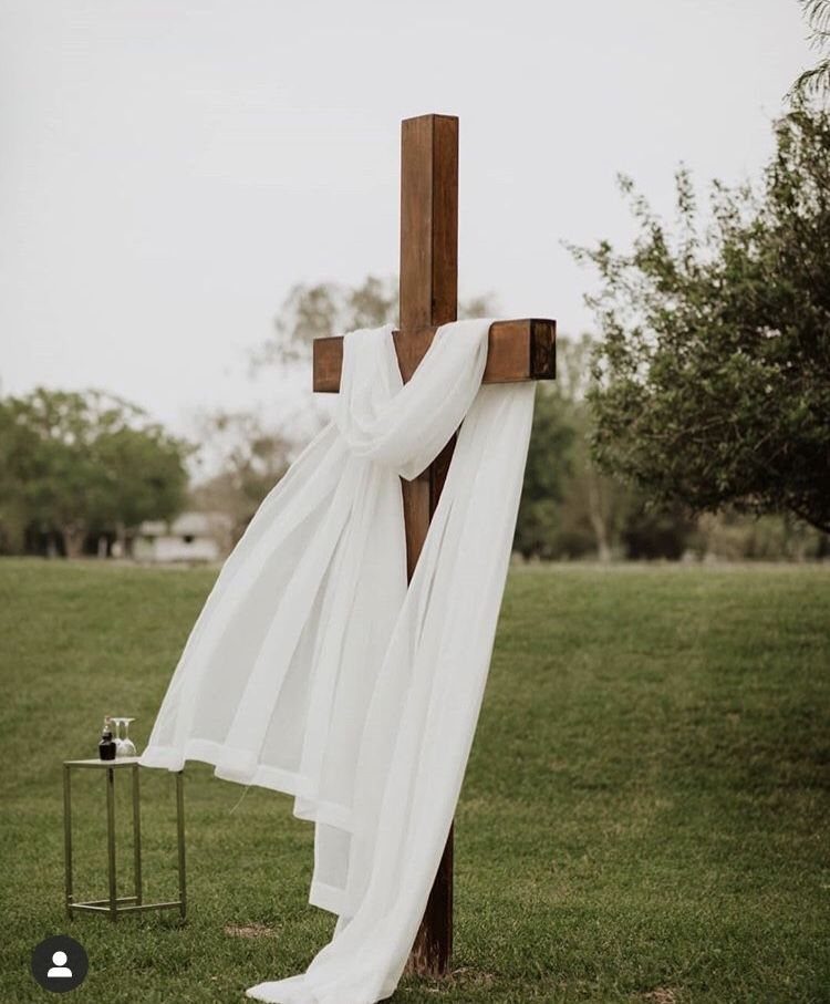 A wooden cross draped with flowing white sheer fabric in an outdoor grassy field.