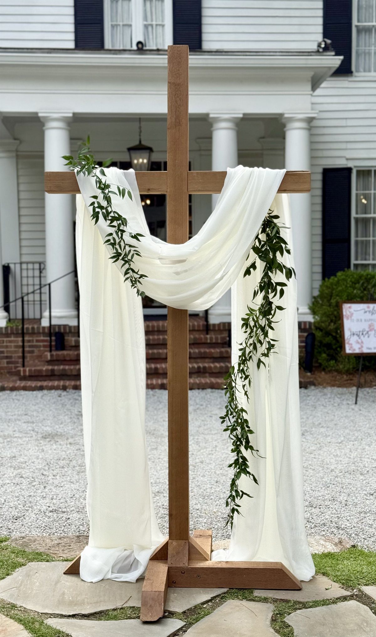 A wooden wedding cross decorated with white draped fabric and green leafy vines outdoors.