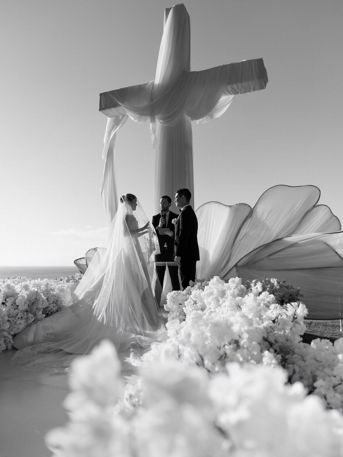 A large cross draped in sheer white fabric stands behind a bride and groom at an outdoor wedding.