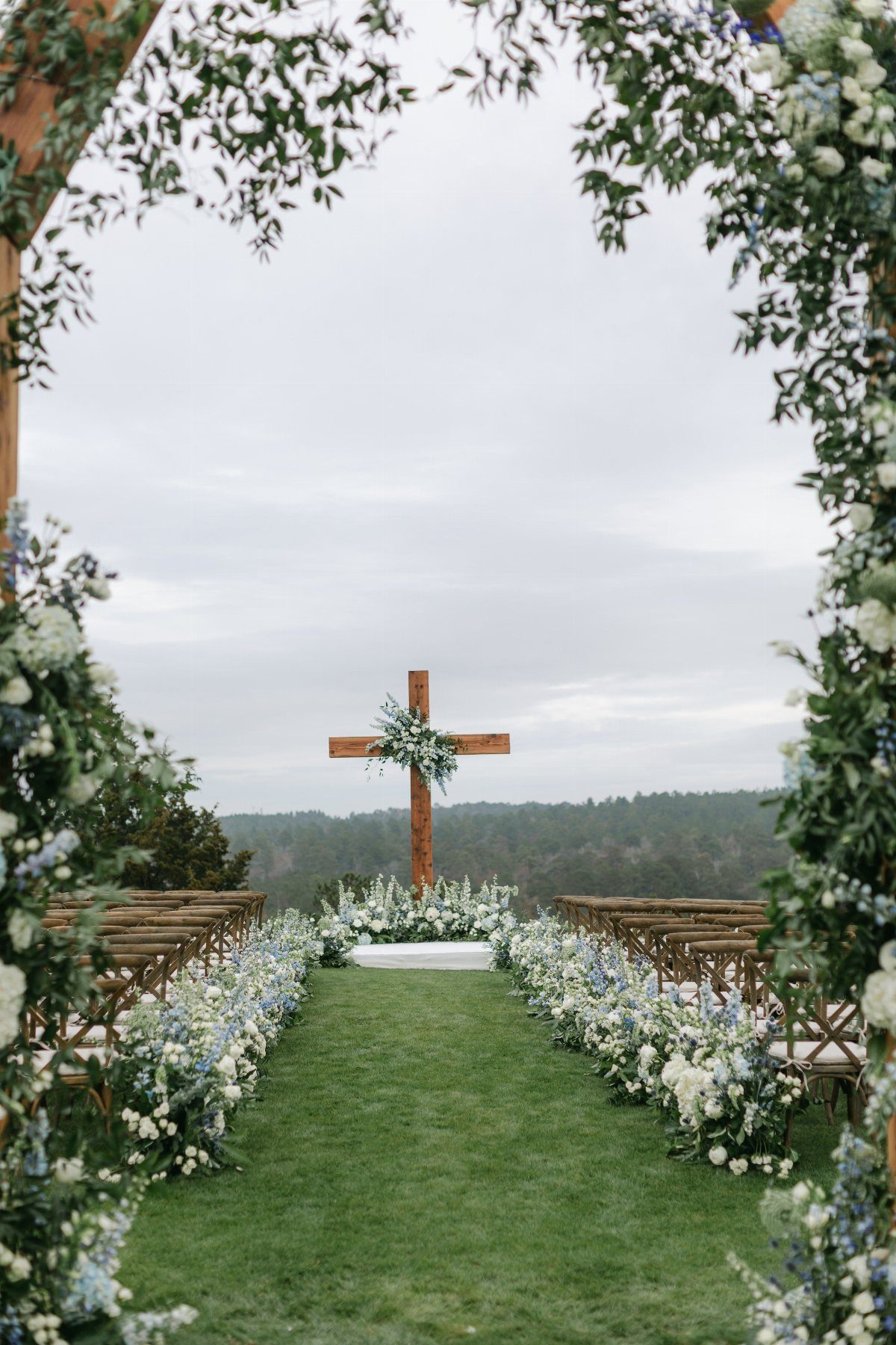 A wooden cross decorated with flowers stands at the end of a grassy wedding ceremony aisle.
