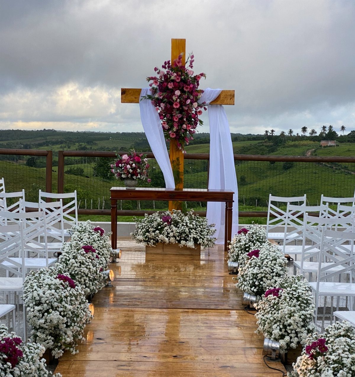 A wooden cross adorned with pink flowers and white fabric stands at an outdoor wedding ceremony altar.