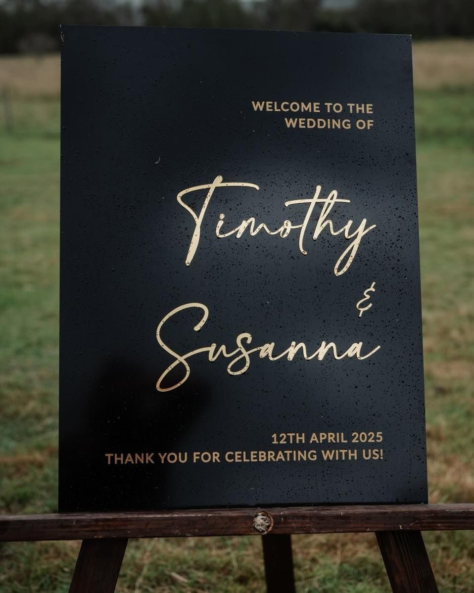 Black wedding welcome sign with gold calligraphy standing on a wooden easel outdoors.
