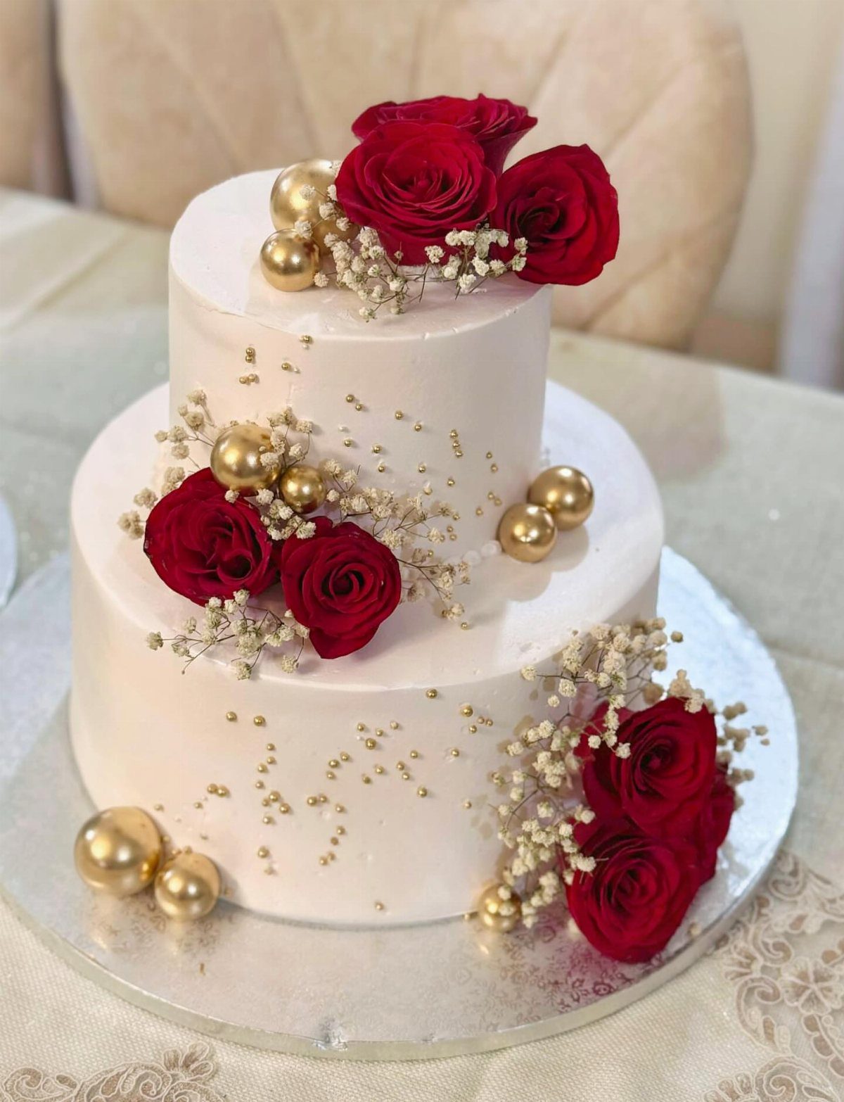Two-tier white cake decorated with red roses, baby's breath, and gold metallic spheres.
