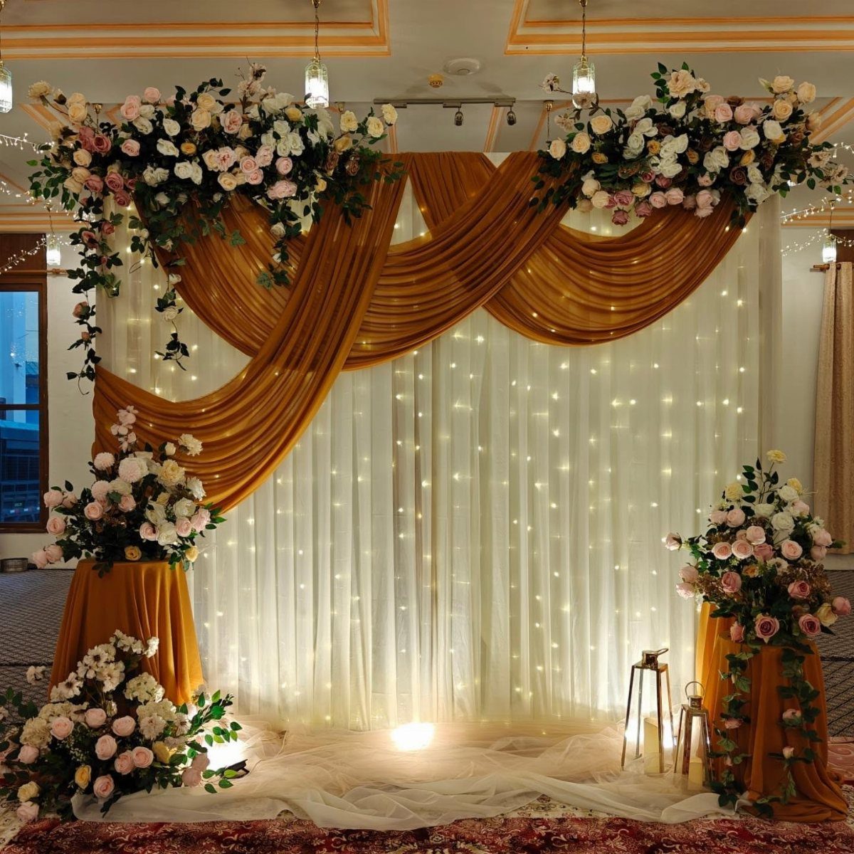 Gold fabric swags draped over white fairy light curtains with large rose floral arrangements.
