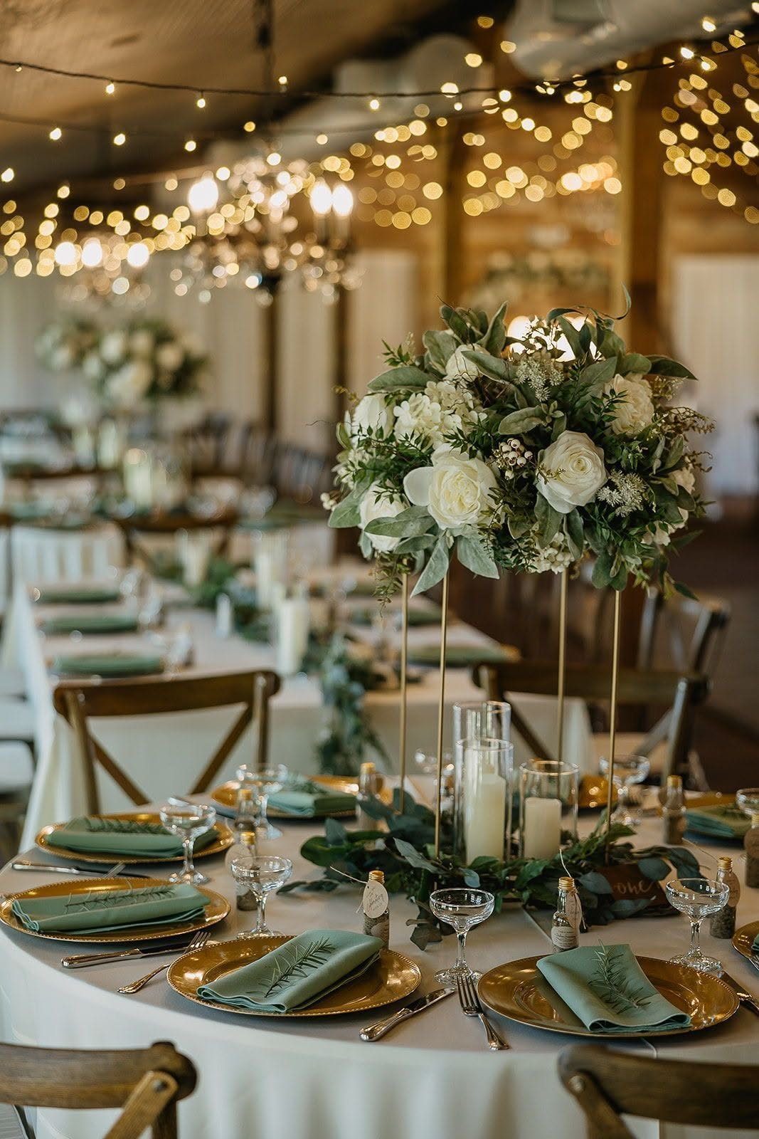 Wedding table setting with white rose and green foliage centerpieces, gold chargers, and sage green napkins.