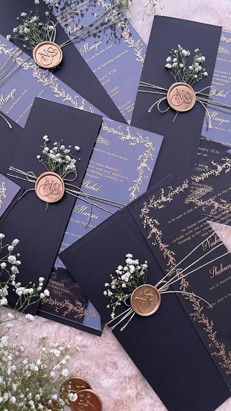 Black wedding invitations with gold calligraphy, twine, baby's breath, and gold wax seals scattered on a surface.