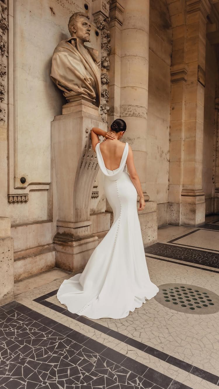 Wedding Dress Backs: 10 Styles That Turn the Exit Into a Moment 9 Woman in a white wedding dress with a draped cowl back standing by stone architecture.
