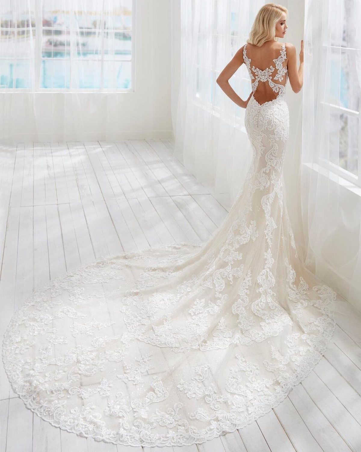 Wedding Dress Backs: 10 Styles That Turn the Exit Into a Moment 4 Woman in a white lace wedding dress with a long train standing in a bright room.