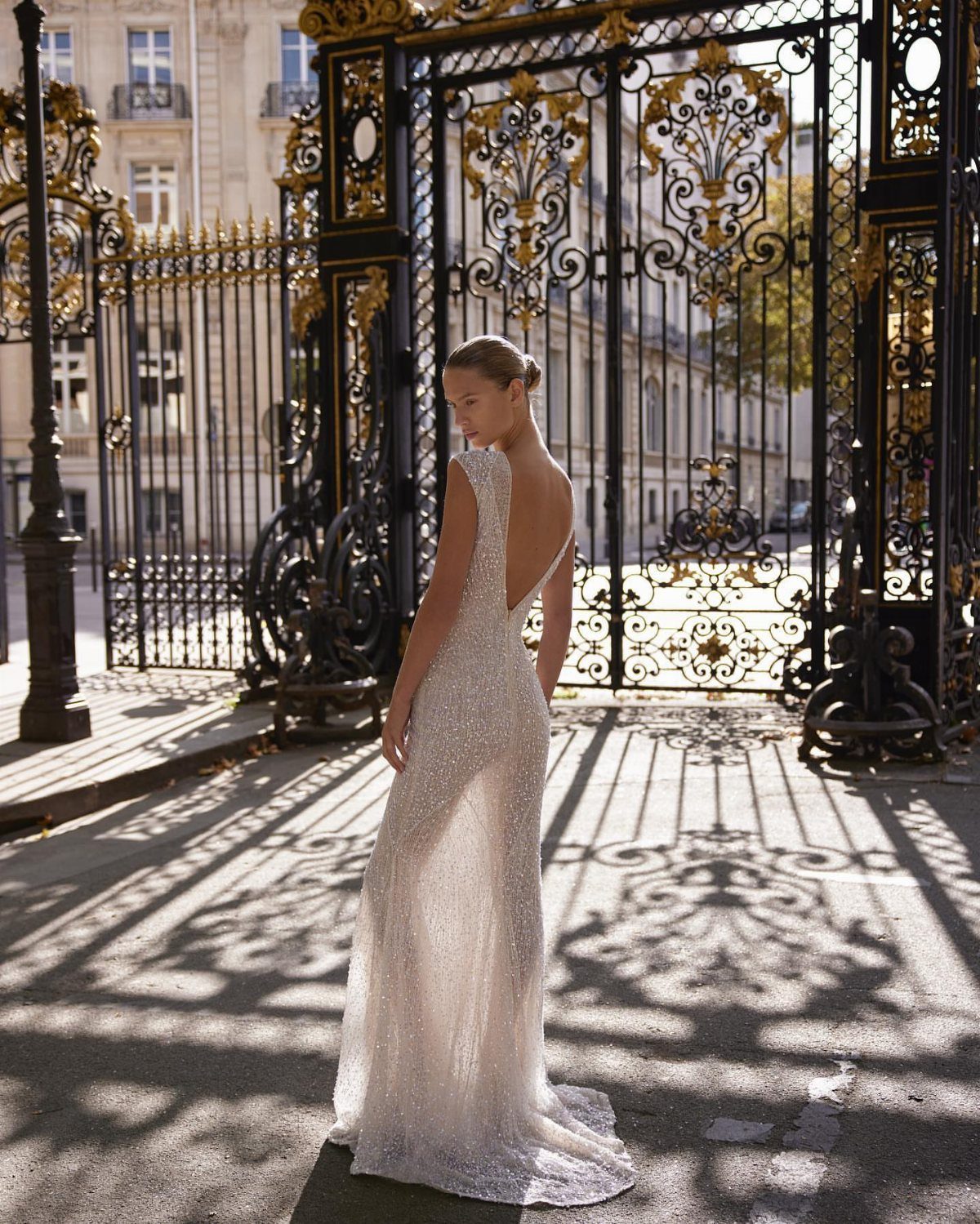 Wedding Dress Backs: 10 Styles That Turn the Exit Into a Moment 7 Woman in a shimmering white wedding dress with a deep low back standing before a golden gate.