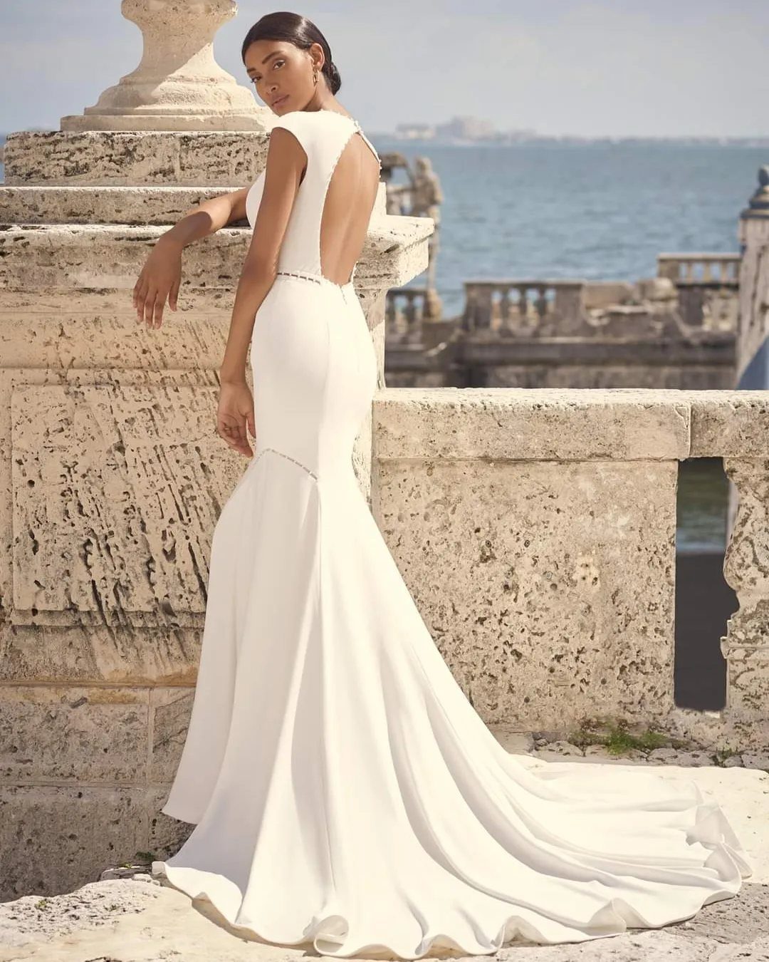 Wedding Dress Backs: 10 Styles That Turn the Exit Into a Moment 6 Woman in a white mermaid wedding dress with an open back standing by a stone wall.