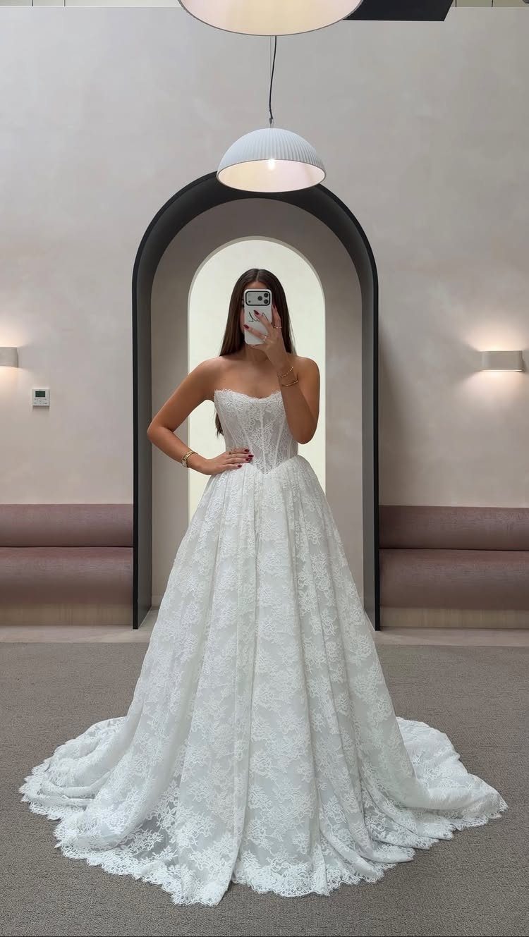 A woman in a white lace strapless ball gown wedding dress posing for a mirror selfie.