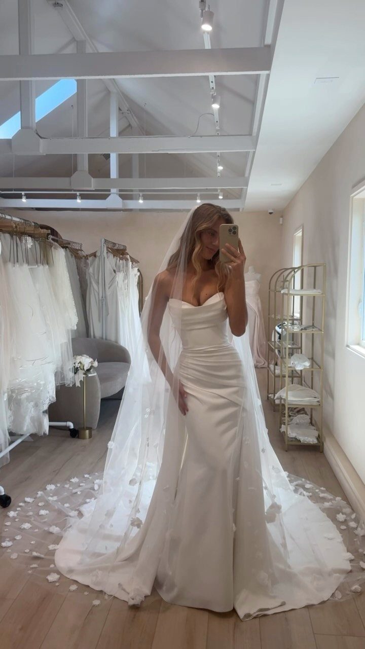 Woman in a white strapless wedding dress taking a mirror selfie in a bridal shop.