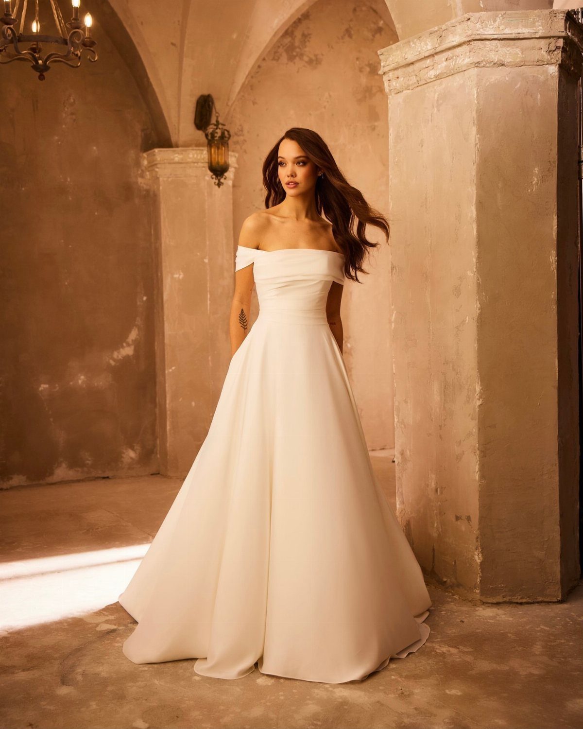 A woman in a white off-the-shoulder satin wedding dress standing in a rustic stone room.