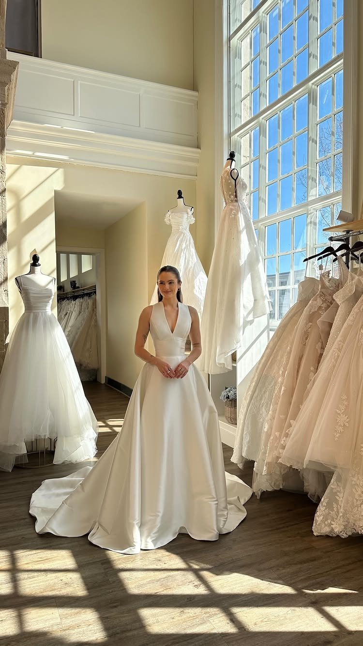 A woman wearing a white satin wedding dress standing in a bright bridal boutique.