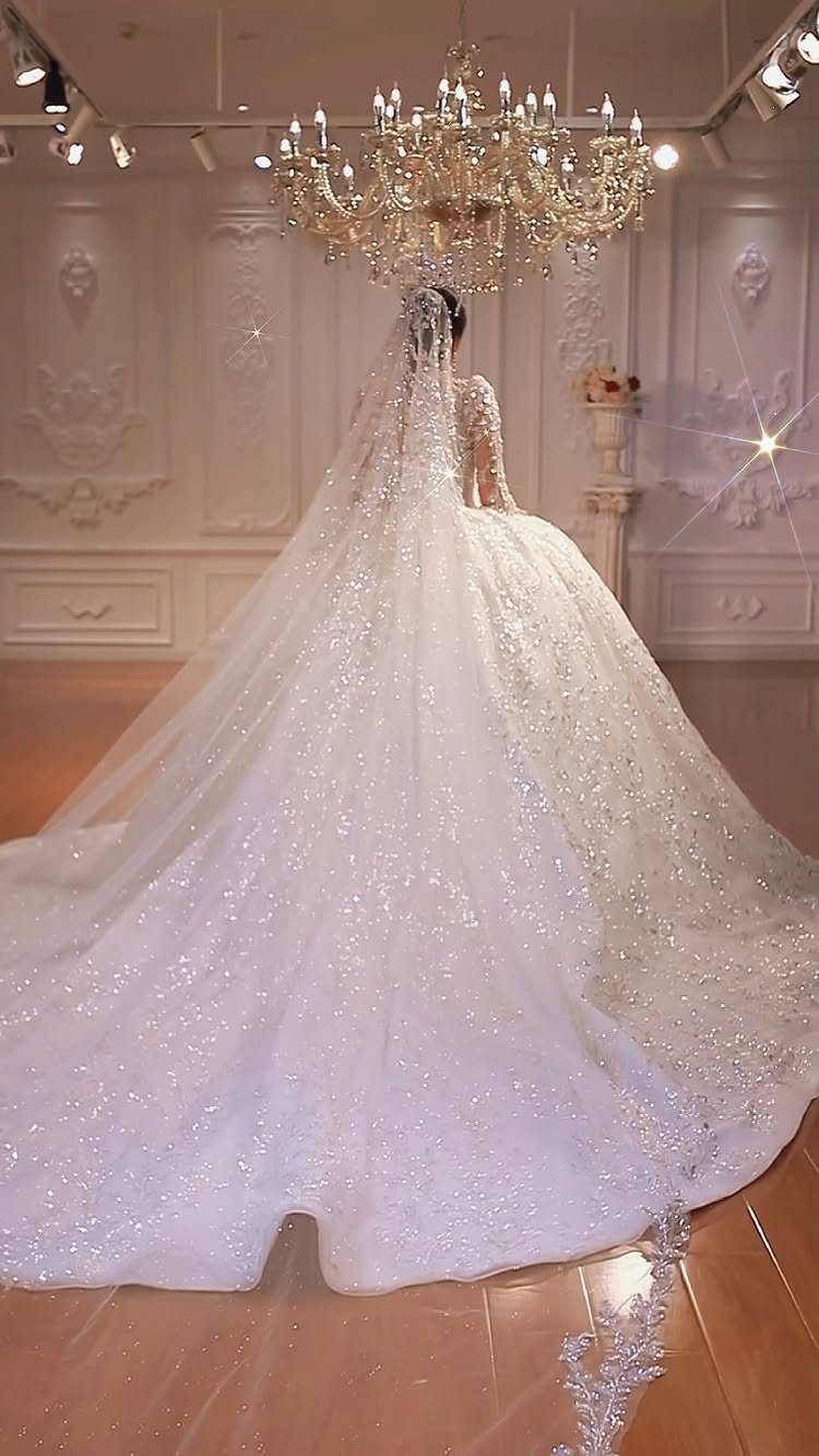 A woman in a sparkling white wedding dress with a long train stands under a chandelier.