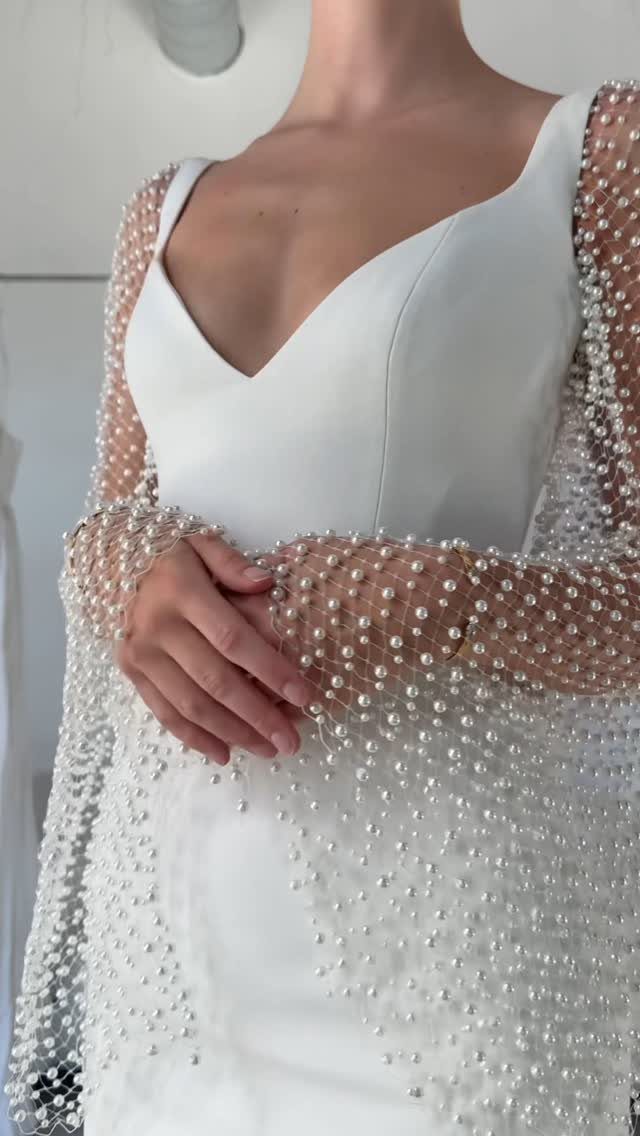 A person wearing a white V-neck wedding dress with a pearl-encrusted sheer cape.