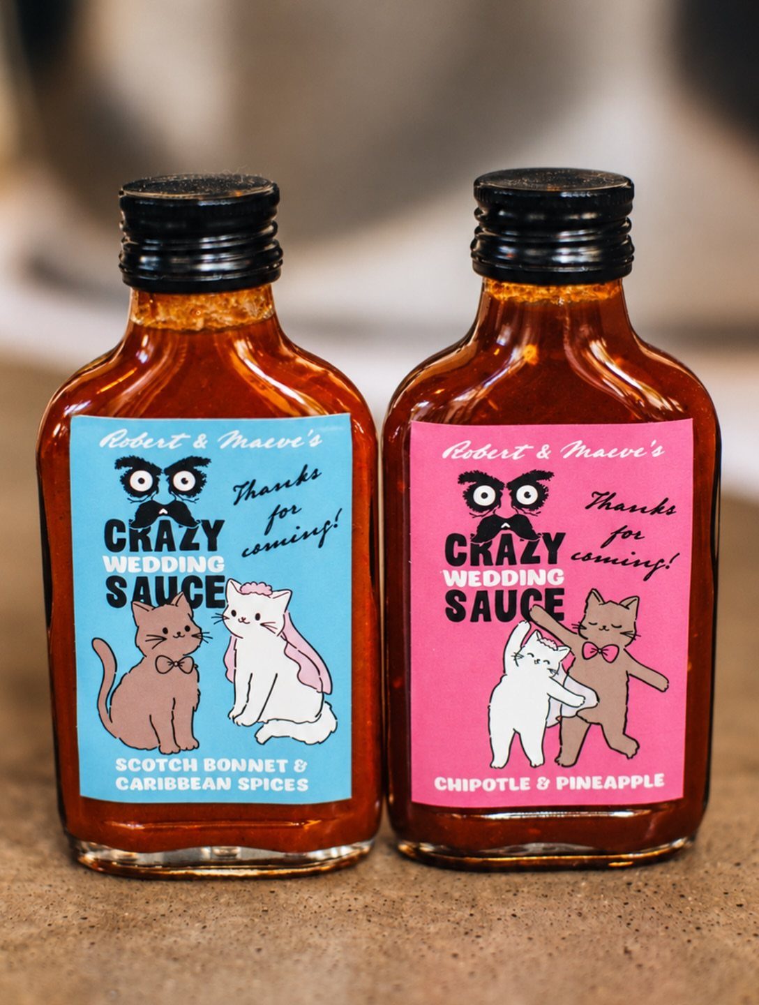 10 Wedding Favors for Guests Ideas: From Plants to Hot Sauce 9 Two small bottles of custom wedding hot sauce with colorful cartoon labels on a stone surface.