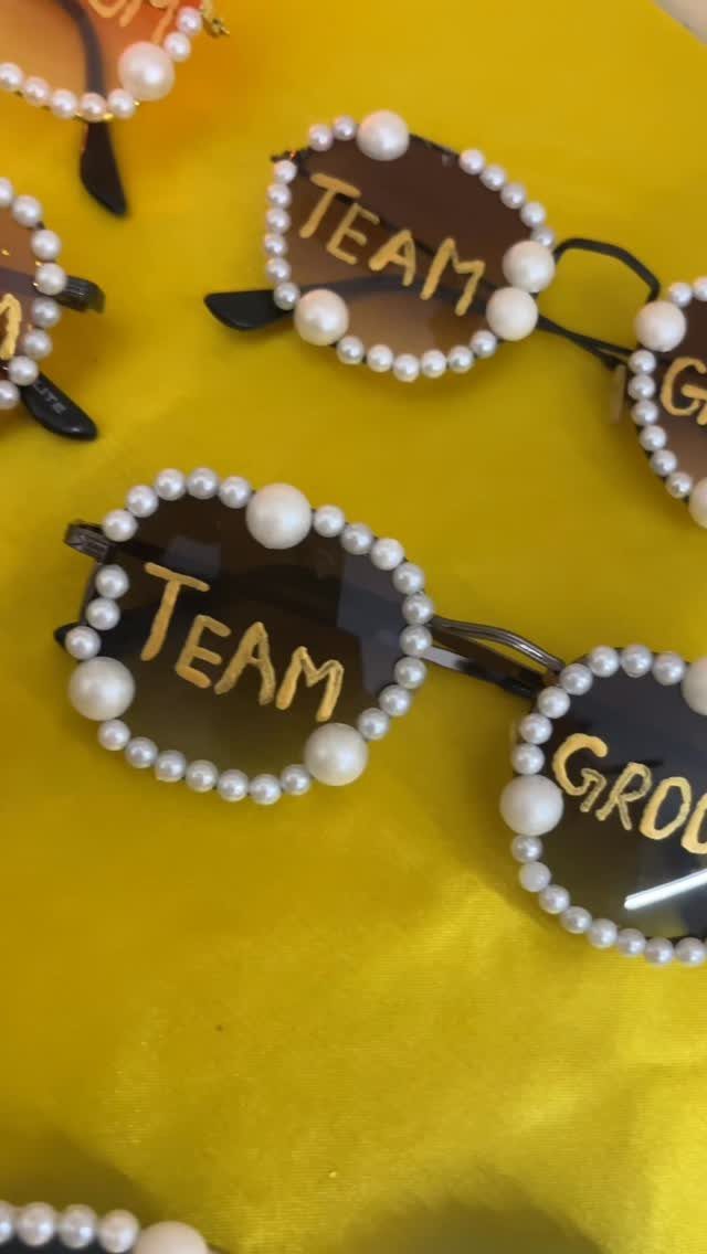 10 Wedding Favors for Guests Ideas: From Plants to Hot Sauce 3 Pearl-decorated sunglasses with gold Team lettering resting on a yellow fabric background.