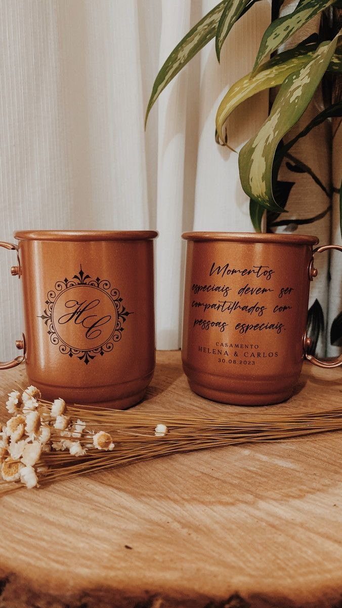 Two copper mugs with black lettering and monograms resting on a wooden table near dried flowers.
