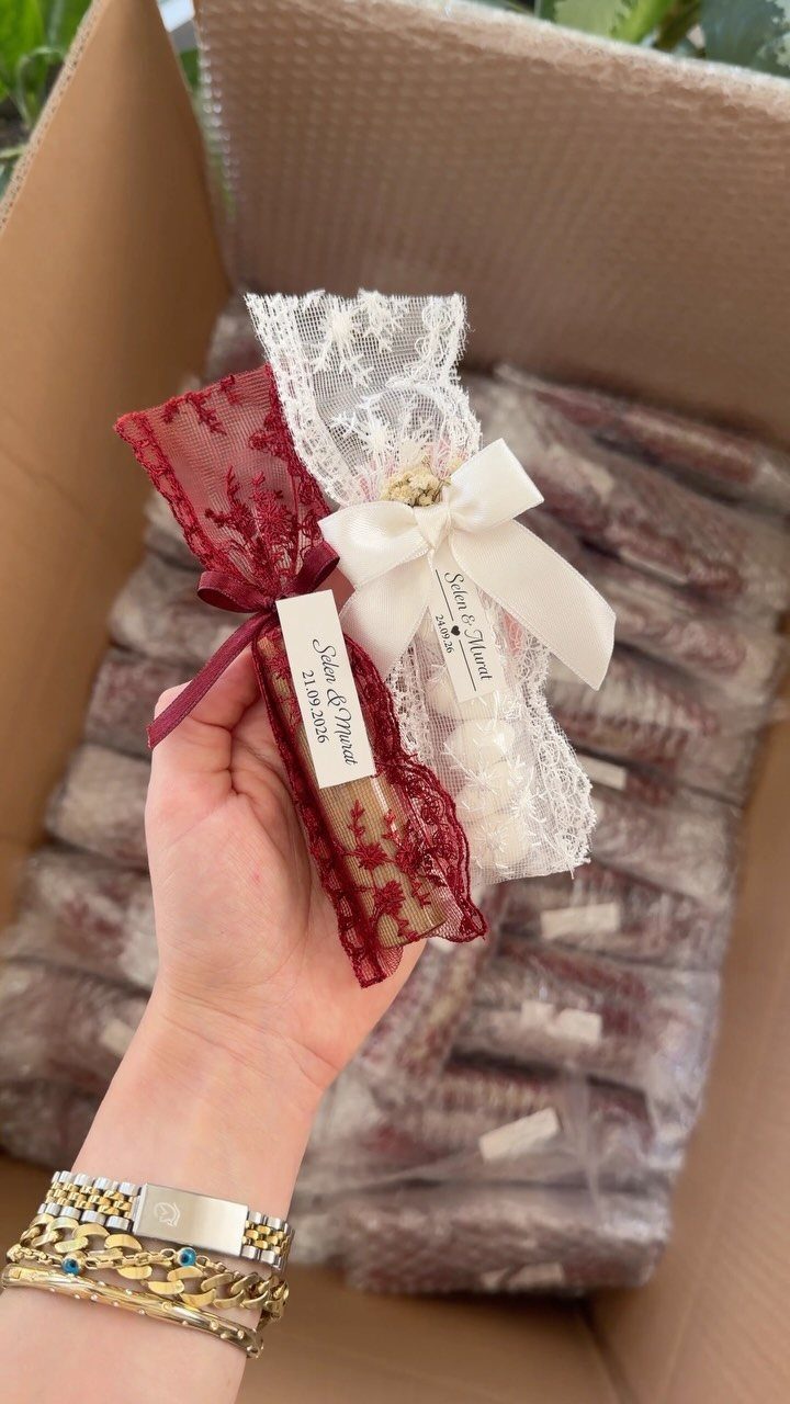 A hand holding two lace fabric favor bags tied with ribbons over a box of gifts.