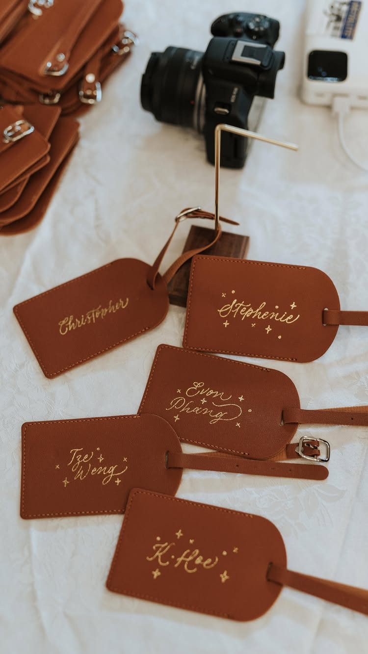 Brown leather luggage tags with gold engraved names scattered on a white surface near a camera.