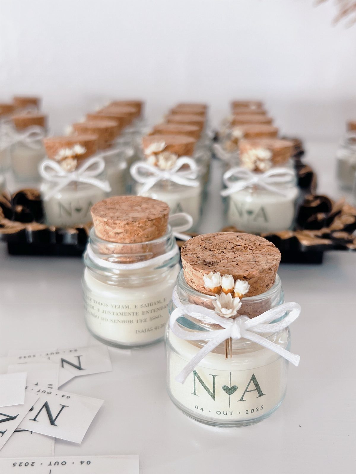 Small glass candle jars with cork lids, white ribbons, and personalized labels on a white surface.