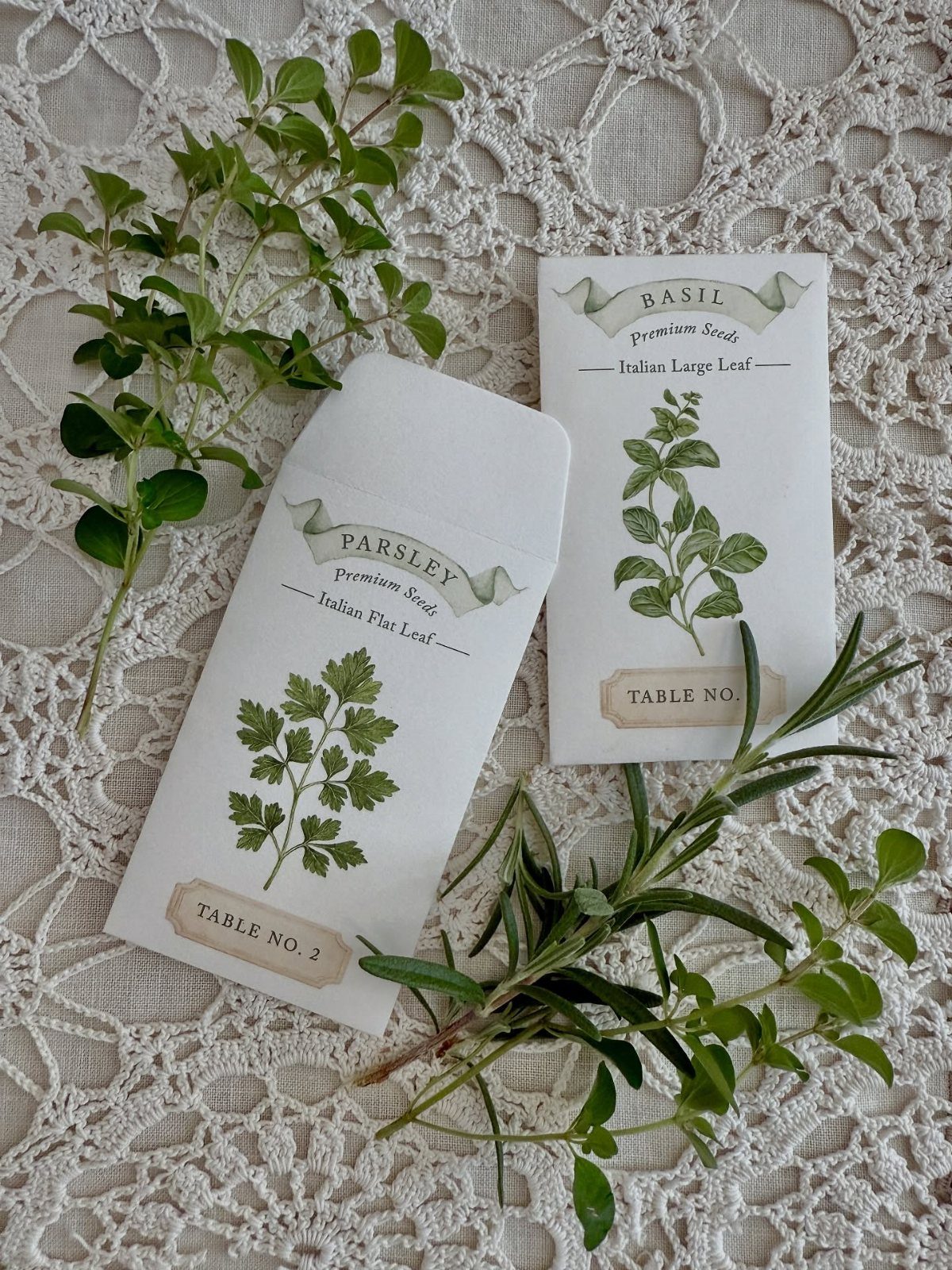 Two botanical seed packets for parsley and basil resting on white lace with fresh herbs.