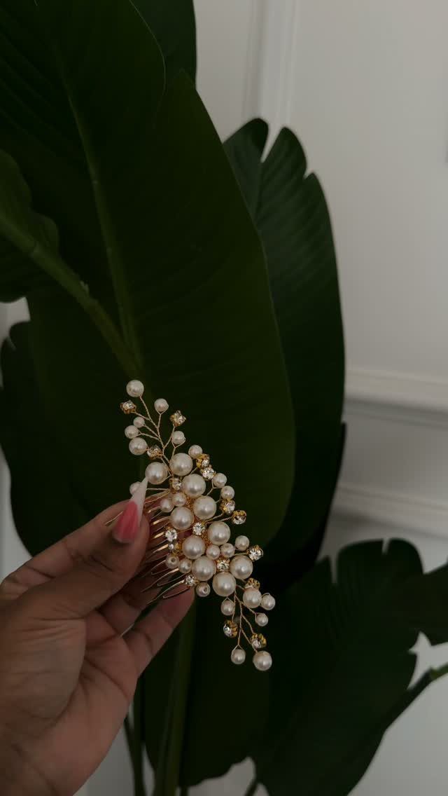 A hand holding a gold hair comb decorated with white pearls and crystals against green leaves.