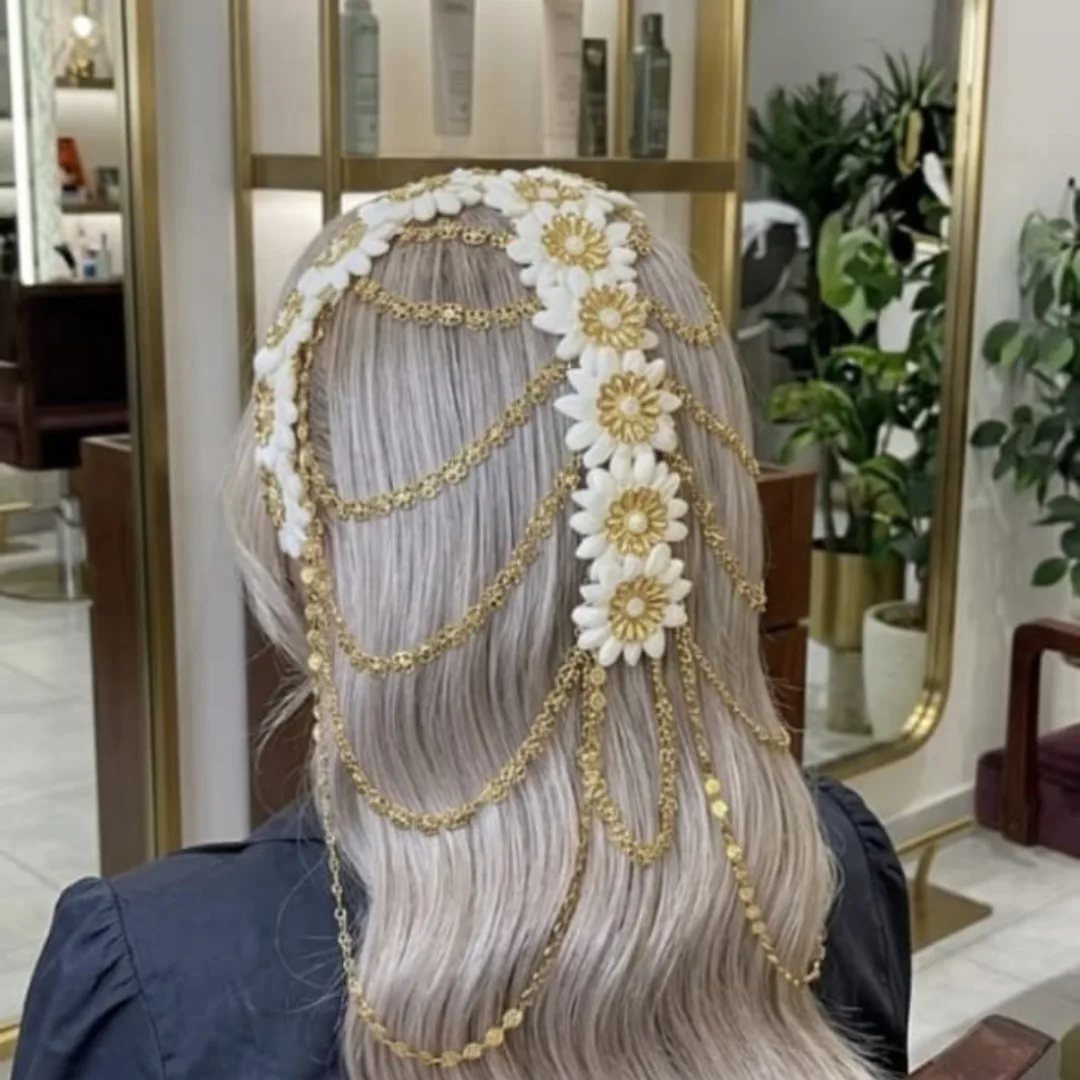 Long blonde wavy hair adorned with a gold floral hair chain and white daisy motifs.
