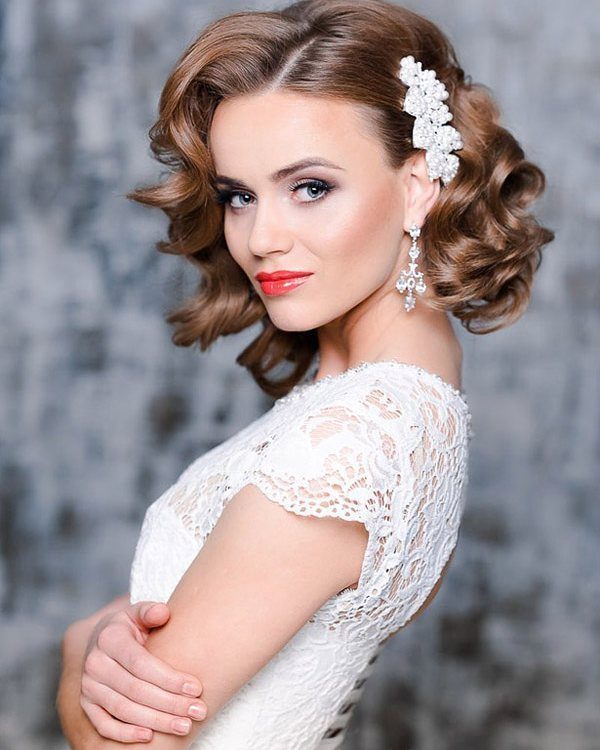 A woman with short wavy brown hair and a white floral hair clip wearing lace.