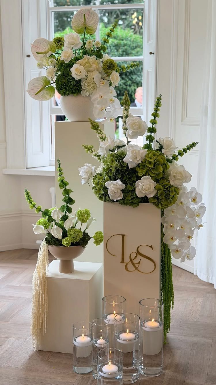 White floral arrangements on cream pedestals with small candles in glass cylinders on the floor.