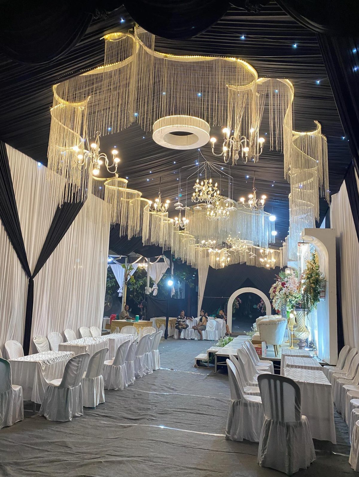 Large event hall featuring white draped tables and multiple hanging chandeliers with long fringe decorations.