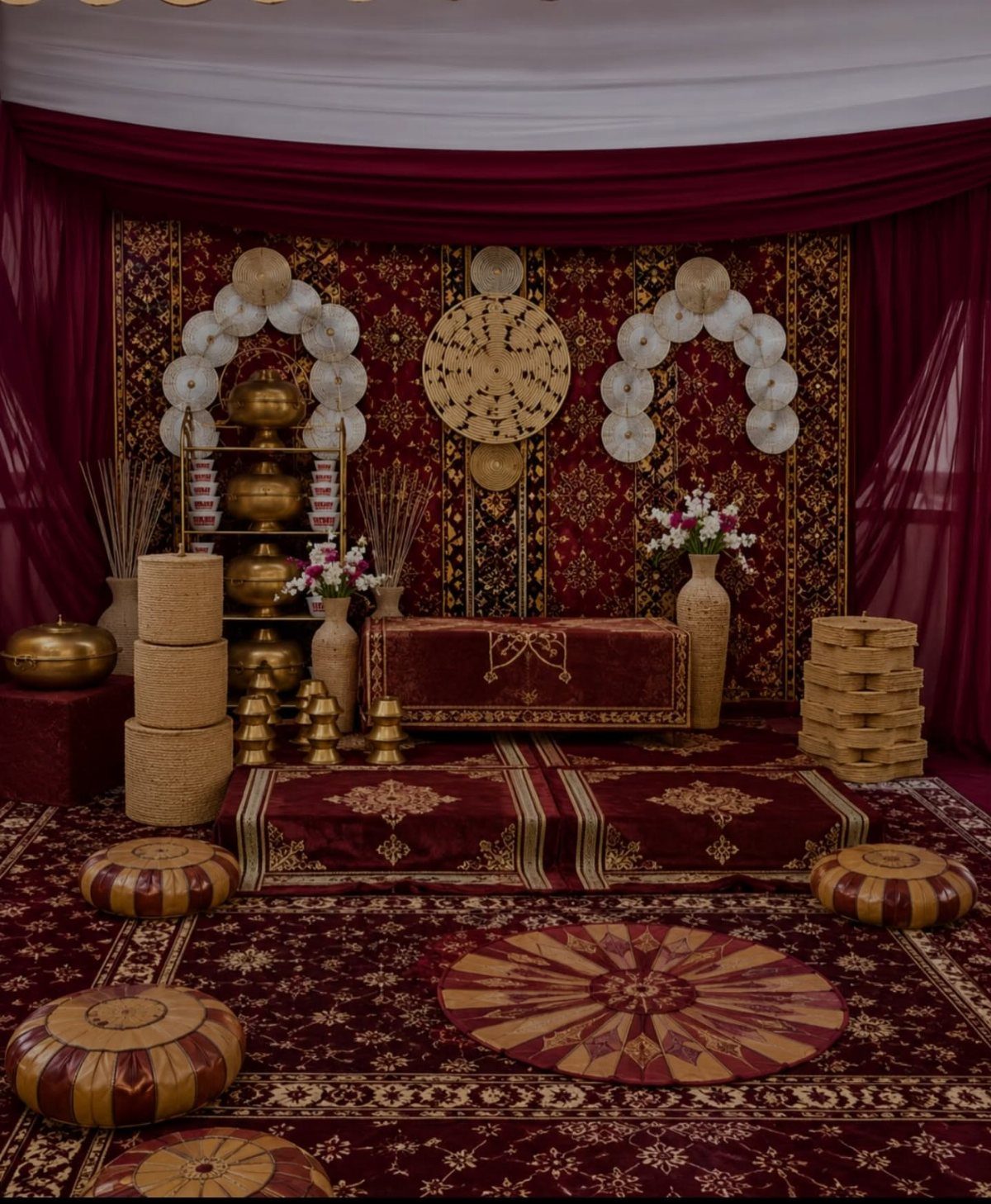 Traditional cultural event decor with red patterned fabrics, golden vessels, and woven baskets on a rug.