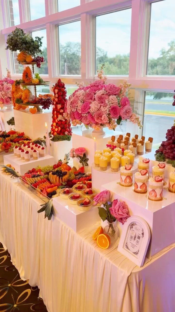 A beautifully arranged dessert table with colorful pastries, fruit, and pink flowers by a window.