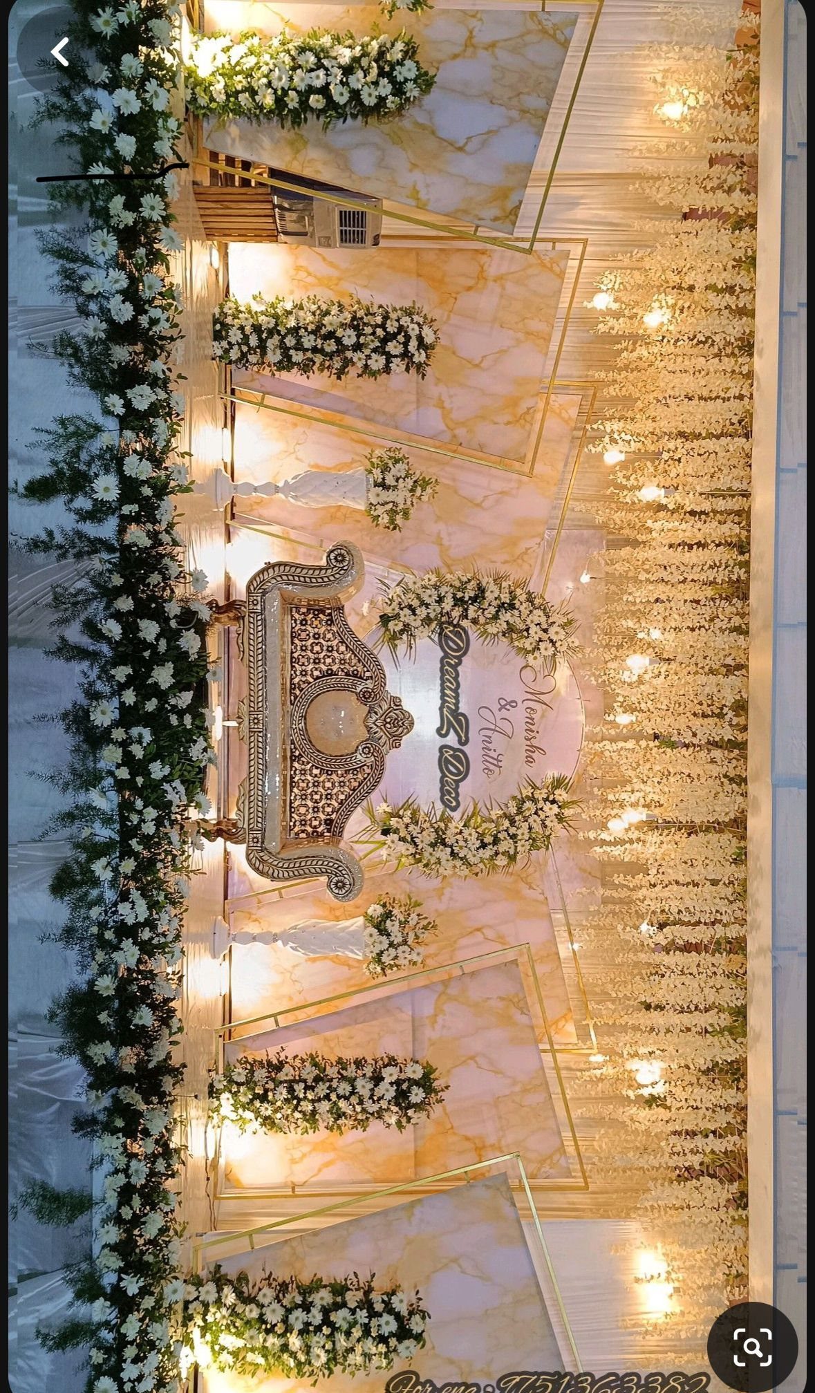 Wedding stage featuring white floral arrangements, gold geometric frames, and hanging fairy lights against marble walls.