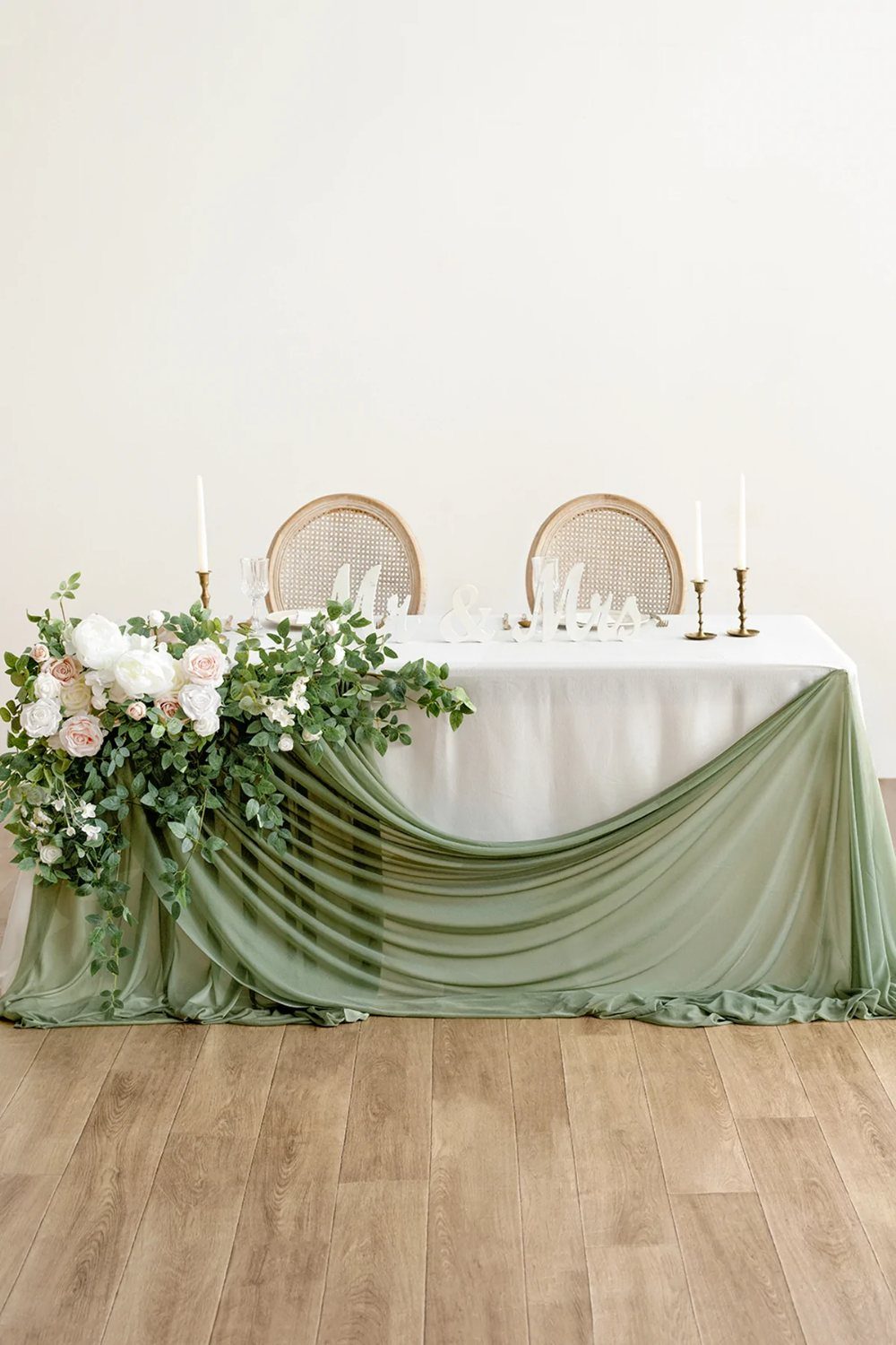 A wedding table decorated with white linens and draped sage green fabric with floral arrangements.