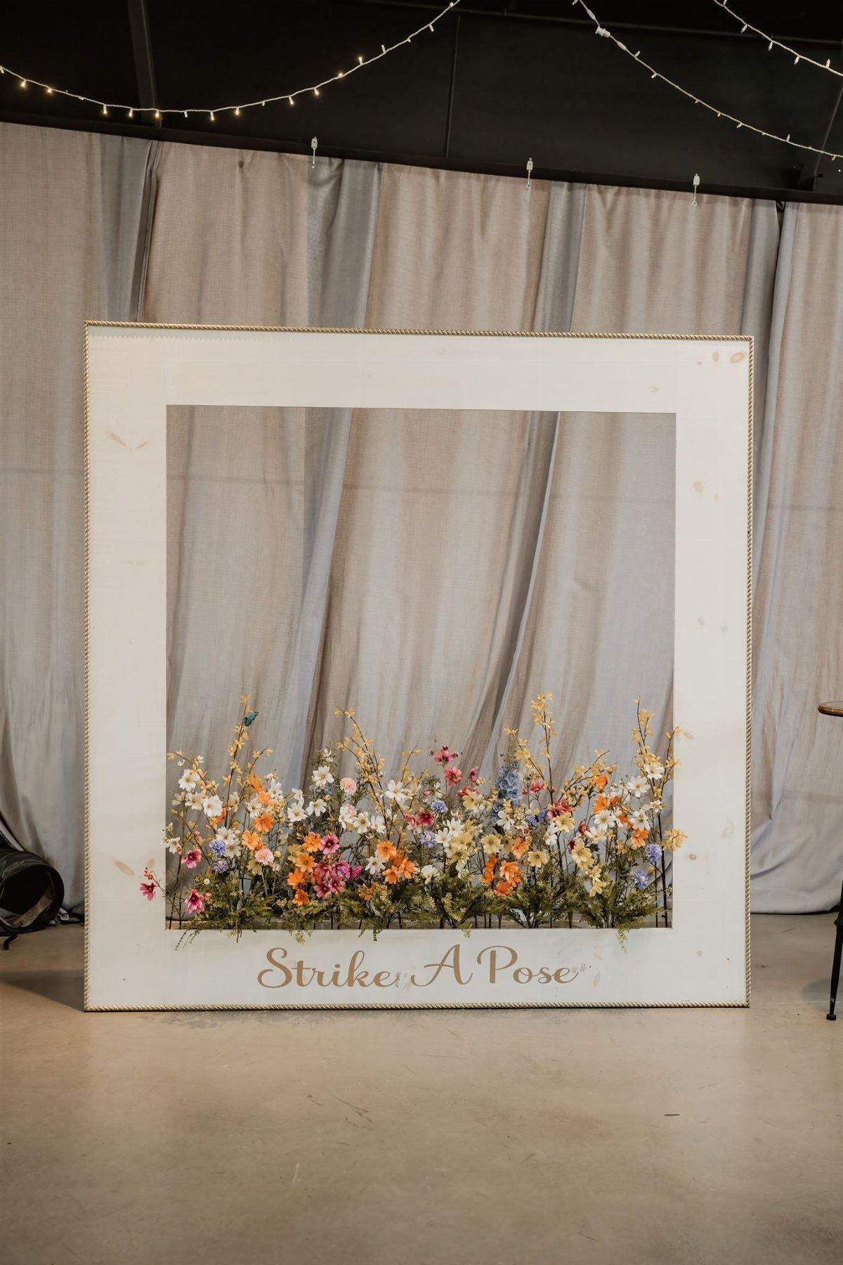 A large white photo booth frame with colorful flowers and text standing before a draped backdrop.