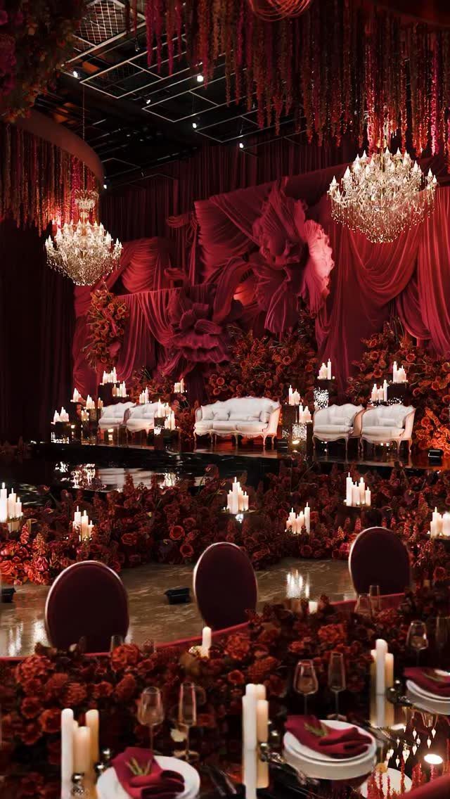 Red floral arrangements and draped fabric decorate a wedding hall with chandeliers and white sofas.