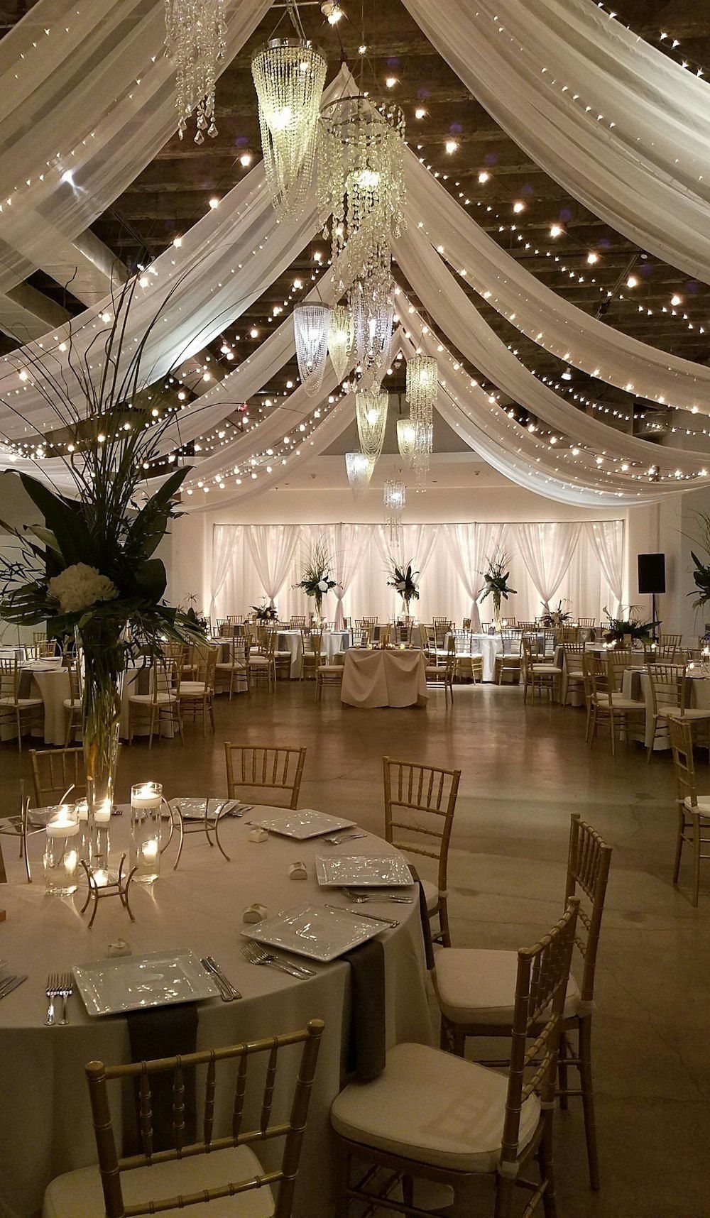 White fabric swags with fairy lights draped across a ceiling above decorated wedding tables.