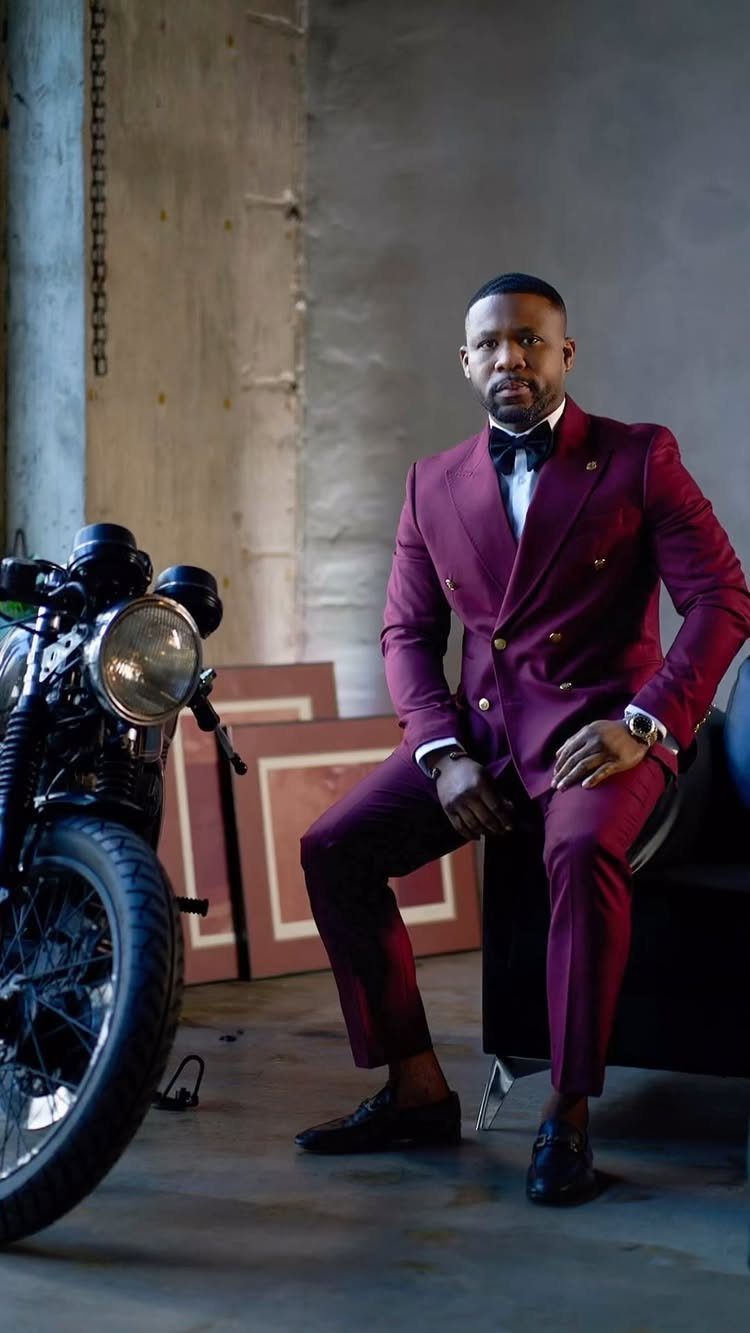 Wedding Suits for Men: 12 Groom Looks Outside Black Tie 11 Man in a burgundy double-breasted suit sitting near a vintage motorcycle in an industrial setting.