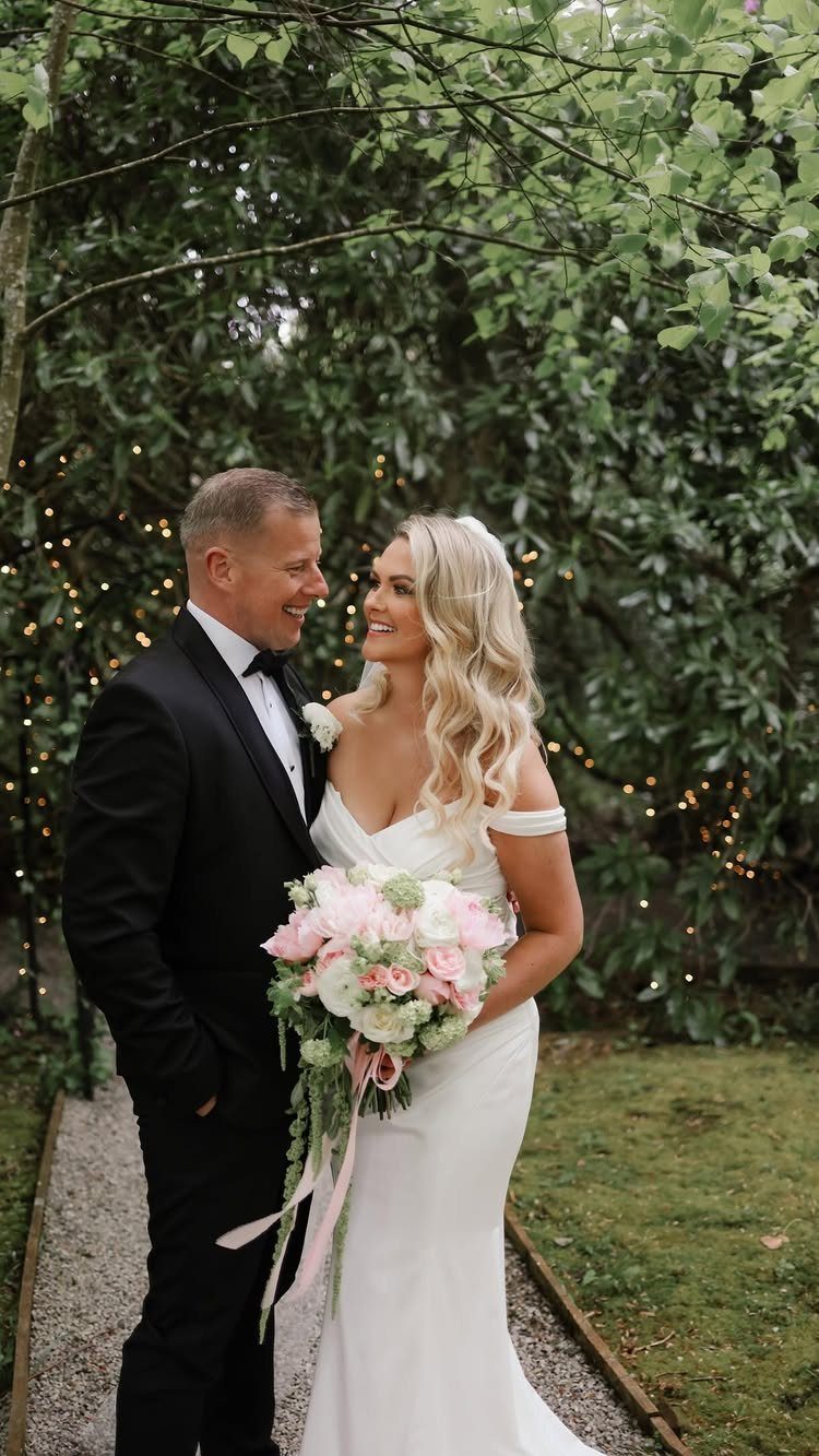 Wedding Suits for Men: 12 Groom Looks Outside Black Tie 3 A groom in a black tuxedo and a bride in white holding flowers outdoors.