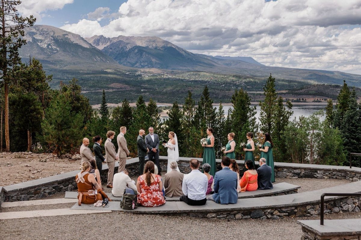Wedding ceremony on a stone amphitheater overlooking a lake and mountains under a cloudy sky.
