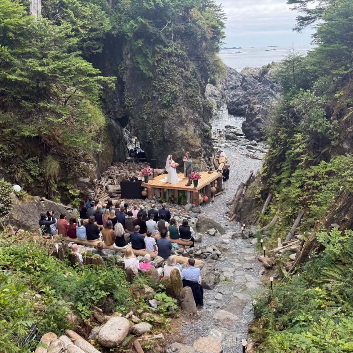 Wedding guests seated on logs watching a ceremony on a wooden platform by a rocky coast.
