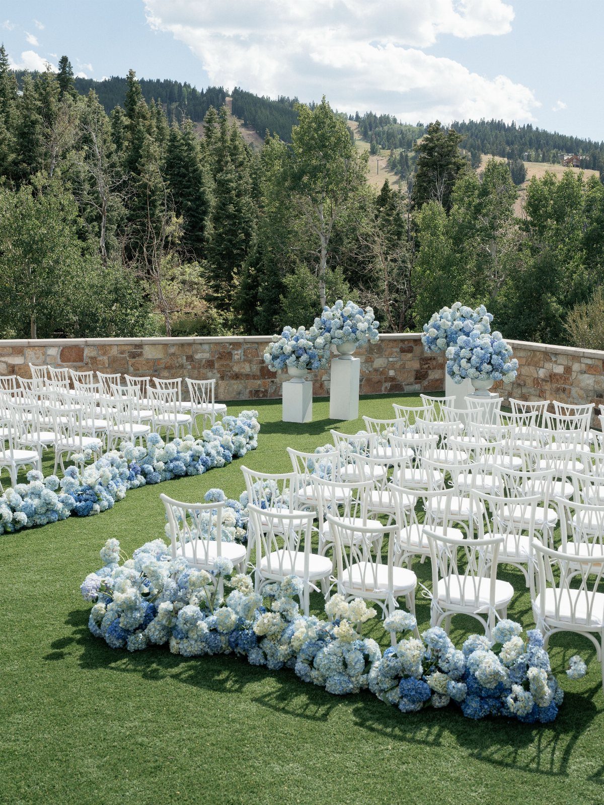 White wedding chairs arranged on green grass with blue hydrangea floral borders near a forest.