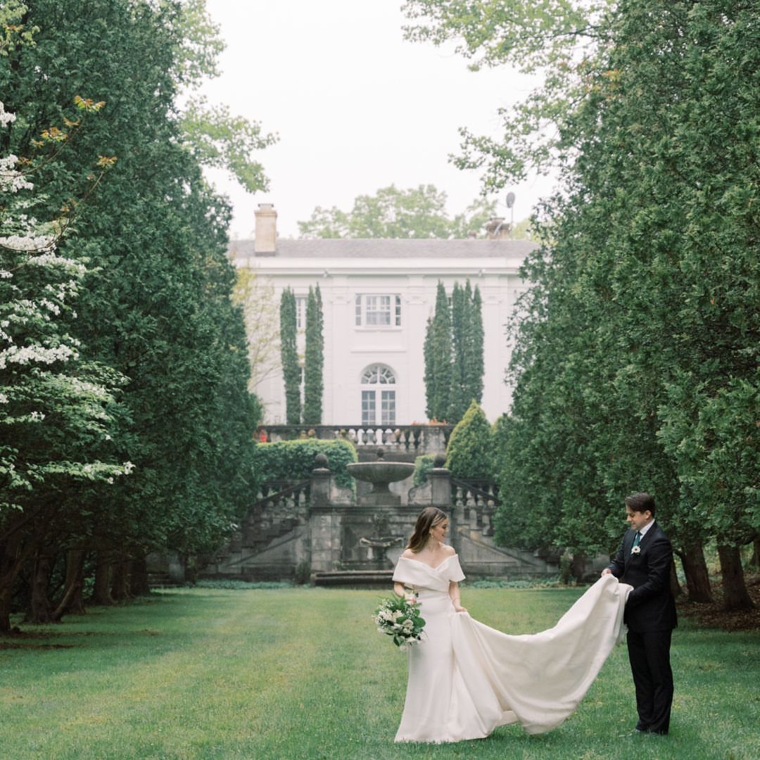A bride and groom pose on a lawn in front of a large white mansion.