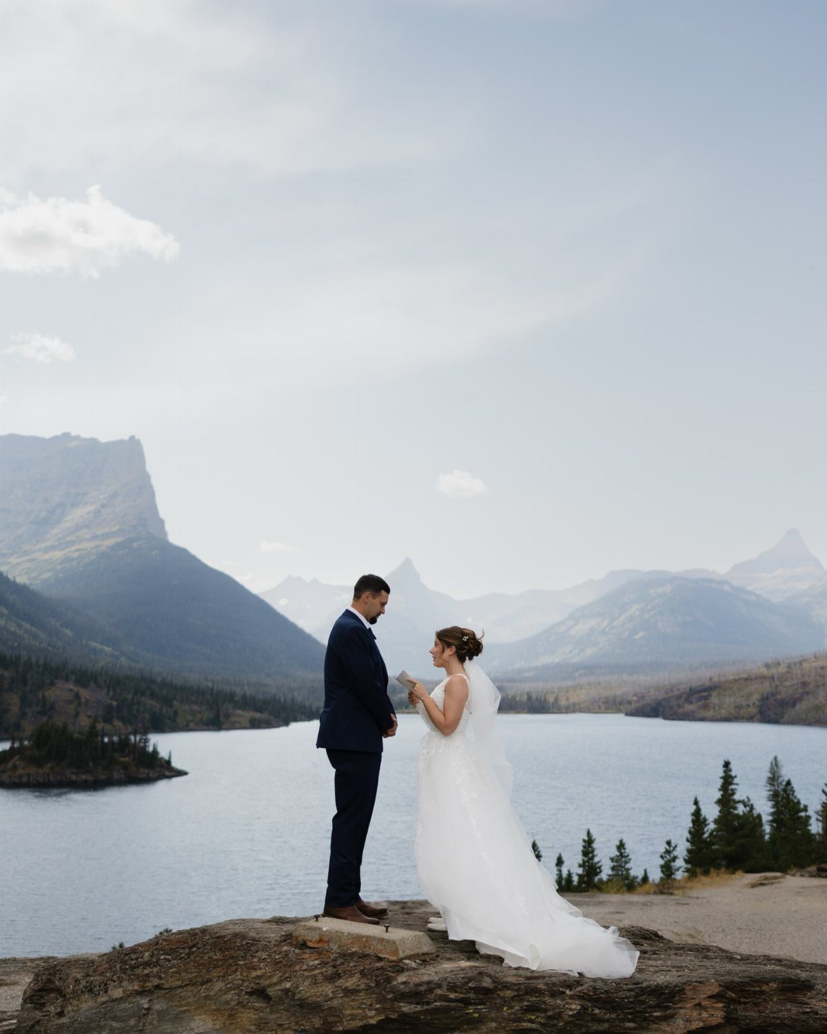 A bride and groom standing on a rocky cliff overlooking a mountain lake.