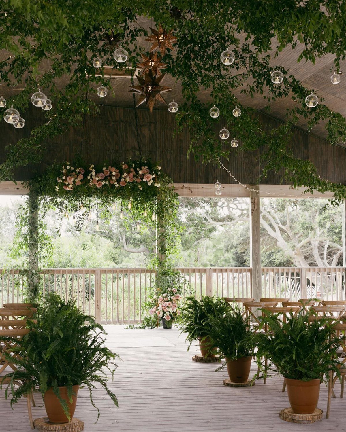 A decorated wooden wedding pavilion with hanging greenery, floral arches, and potted ferns overlooking trees.