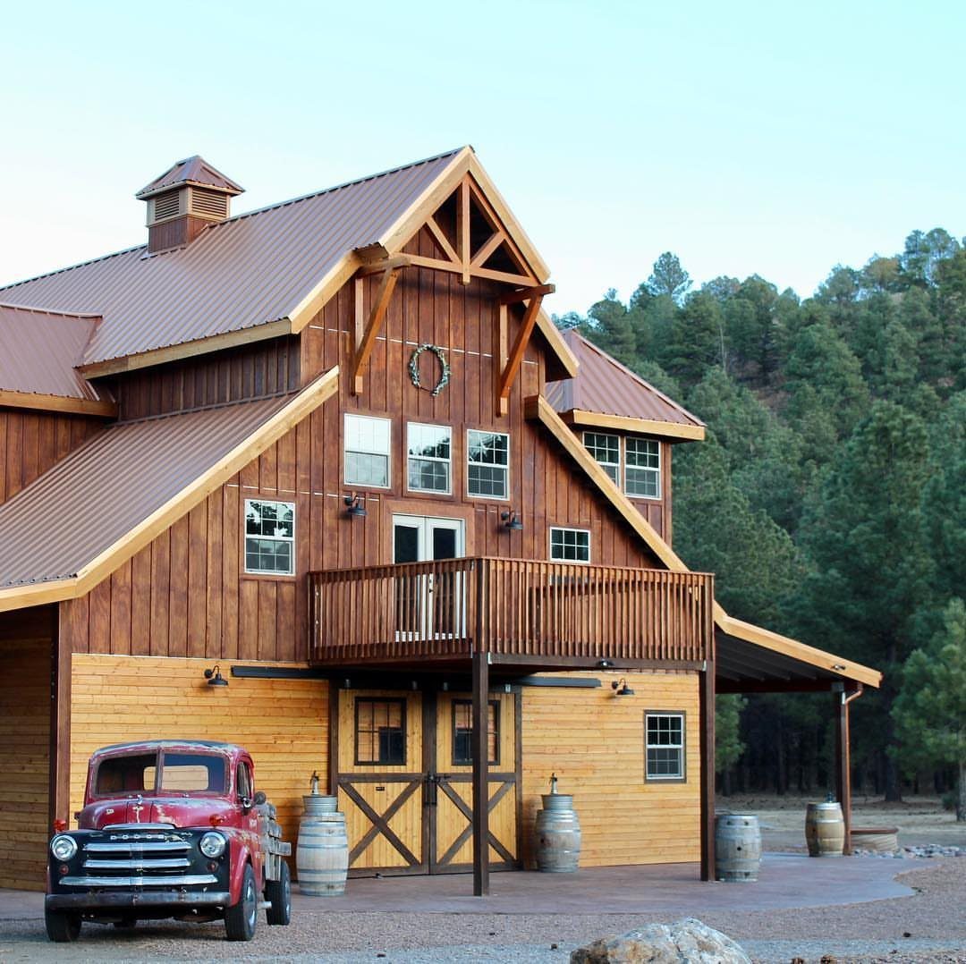 A large wooden rustic barn with a red vintage truck parked in front of a forest.